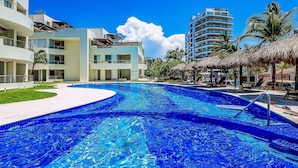 A swimming pool with a curved edge, a thatched-roof structure, and a multi-story building in the background. - Beachfront Resort Condo w/ Pool, Rooftop Lounge & Tropical Ocean Views (Nuevo Vallarta)