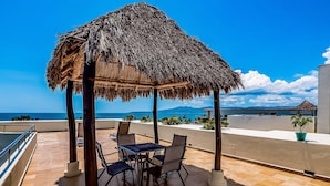 A thatched-roof outdoor seating area with a table and chairs, overlooking a coastal view. - Beachfront Resort Condo w/ Pool, Rooftop Lounge & Tropical Ocean Views (Nuevo Vallarta)