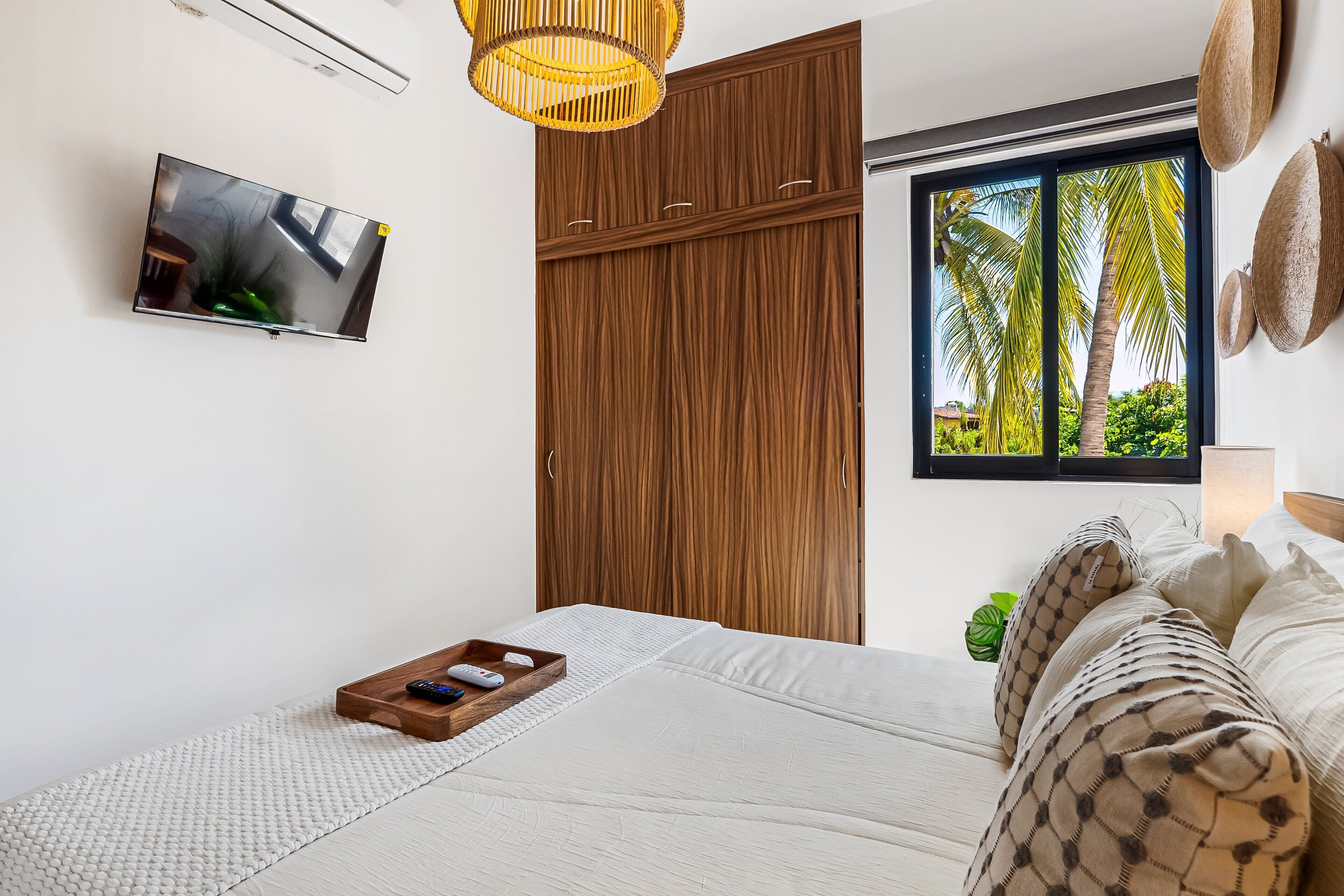 A bedroom with a bed, a TV, a wardrobe, a window with a view of palm trees, and a bedside table with a tray.