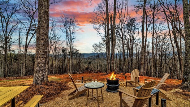 A forested campsite with a fire pit, Adirondack chairs, and a picnic table.