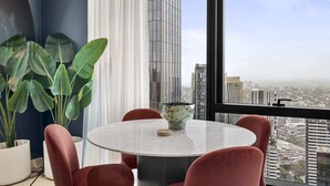 Une pièce moderne avec une table ronde, des chaises rouges, une grande plante en pot et une vue sur le paysage urbain, visible à travers une grande fenêtre.