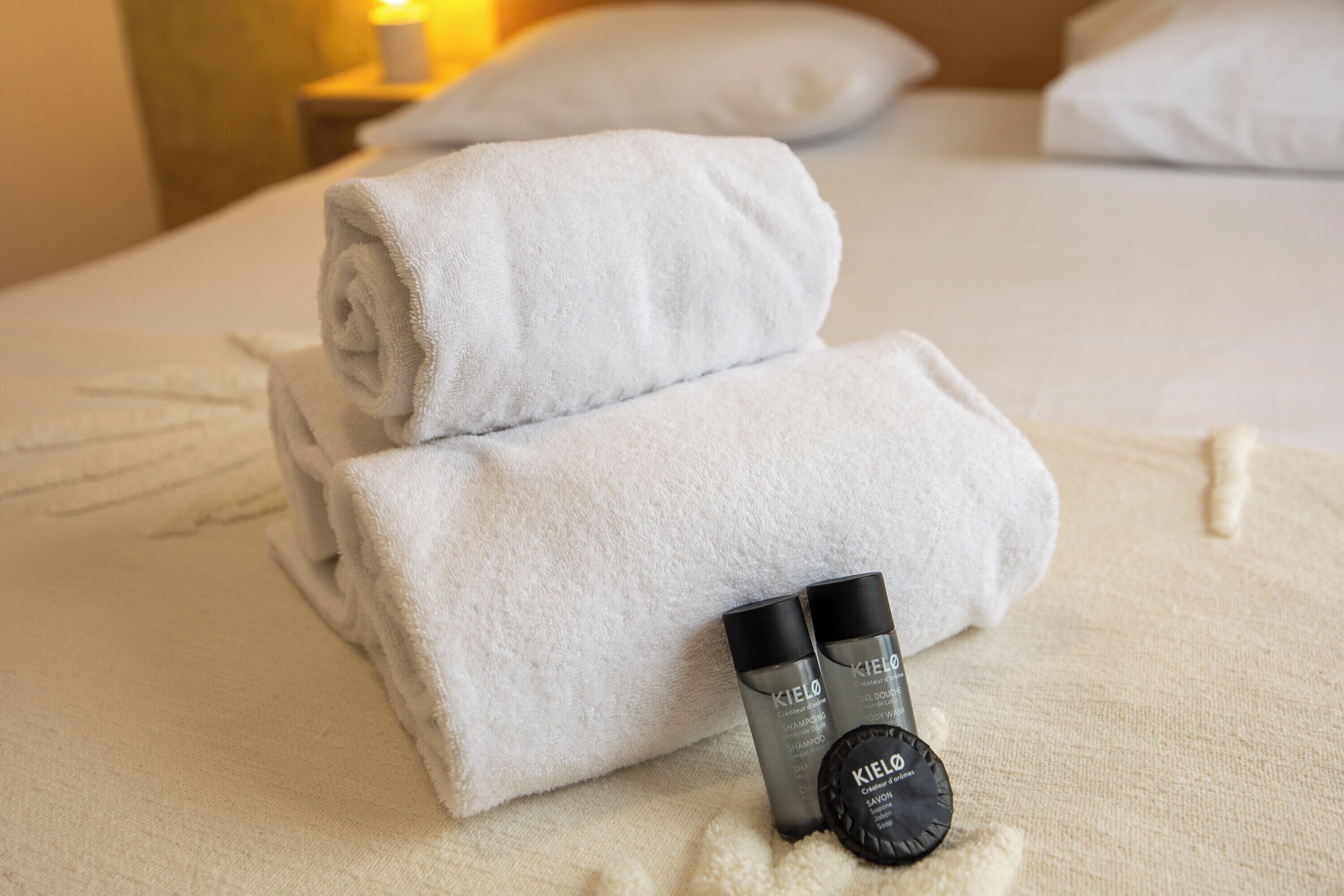 Una cama bien hecha con dos toallas blancas enrolladas y dos botellas de jabón corporal y champú Kiehl's.