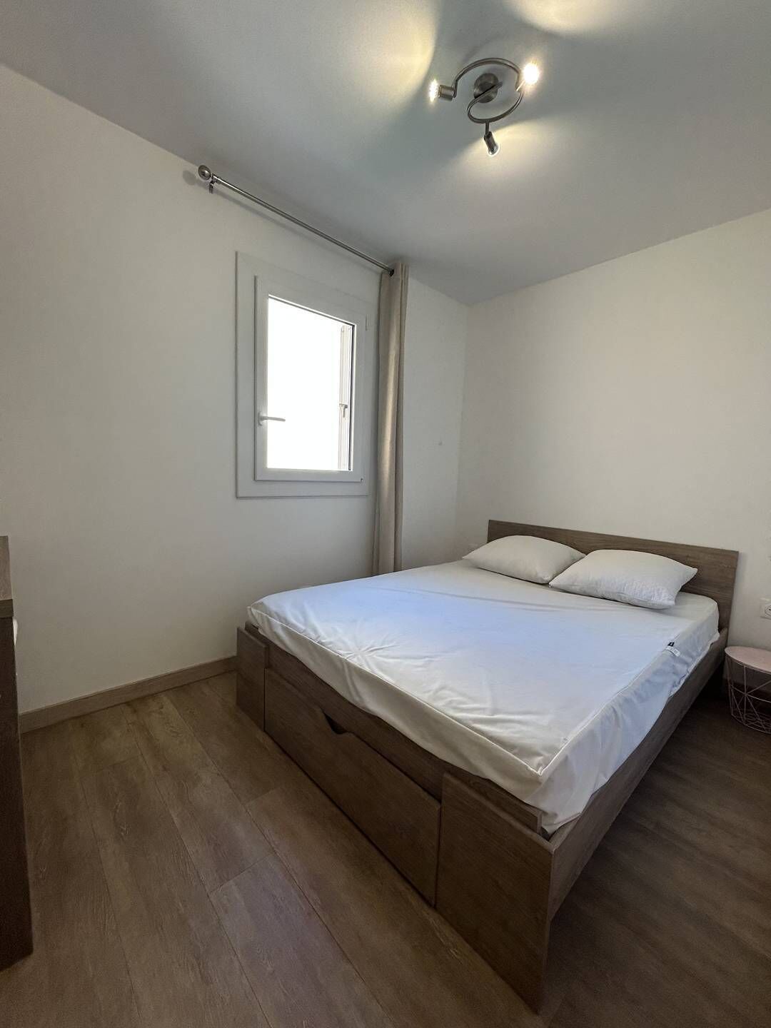 A bedroom with a bed, a window, a ceiling light, and a wooden floor.