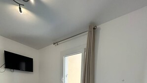 A hotel room with a bed, a desk, a TV, a window with curtains, and a ceiling light. - Lovely 2-room with cabin apartment near the port and shops in Cap d'Agde (LE CAP D AGDE)