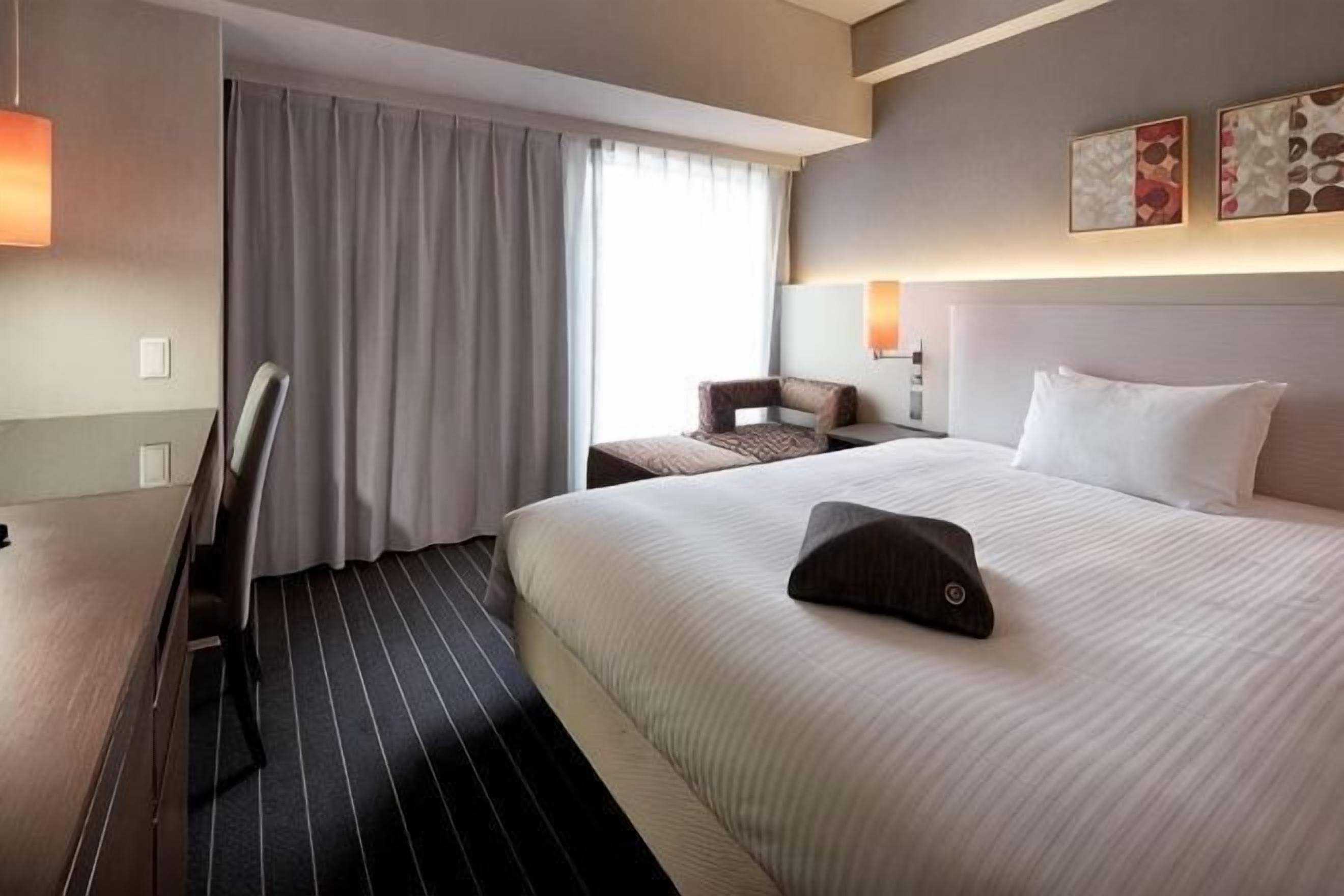 Een hotelkamer met een bed, een bureau, een stoel, een nachtkastje en een raam met gordijnen.