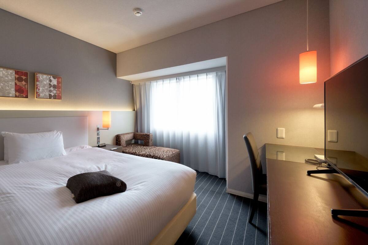Habitación de hotel con una cama grande, un escritorio, una silla, una lámpara y una ventana con cortinas.