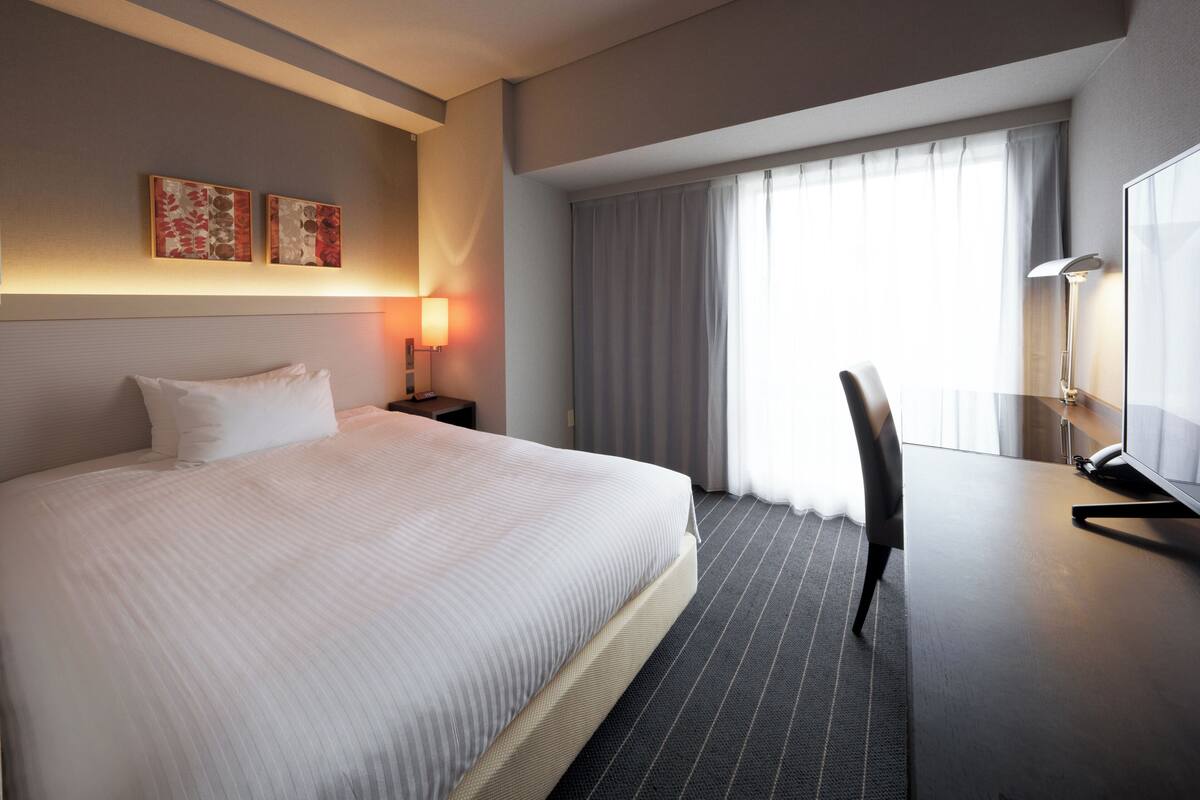 Habitación de hotel con una cama grande, un escritorio con silla, un televisor de pantalla plana y un ventanal con cortinas.