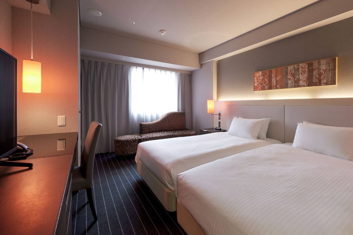 Habitación de hotel con dos camas, un escritorio con silla, un televisor y una ventana con cortinas.