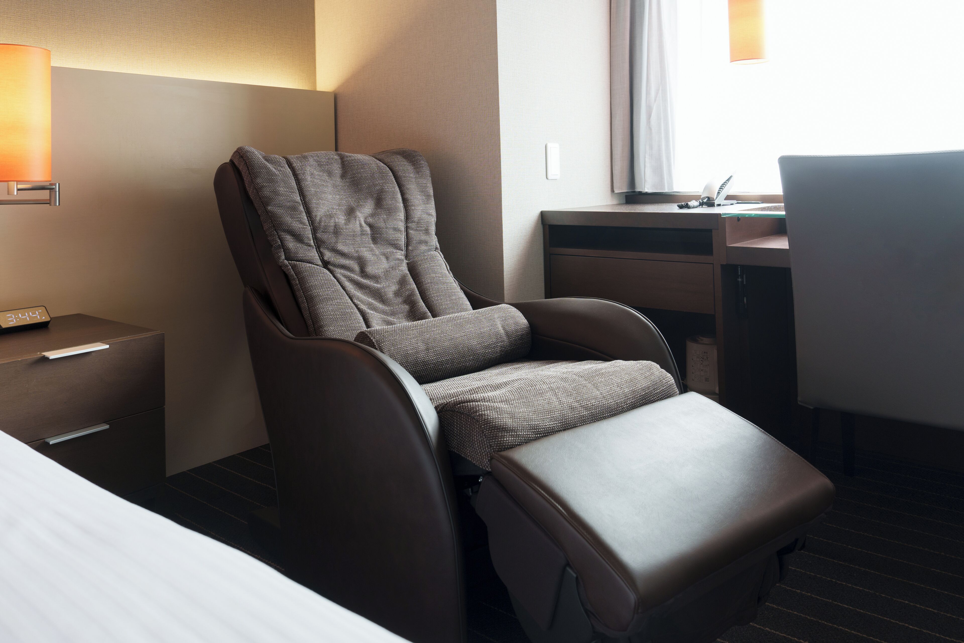 A modern hotel room with a reclining chair, a desk, a lamp, and a bedside table.