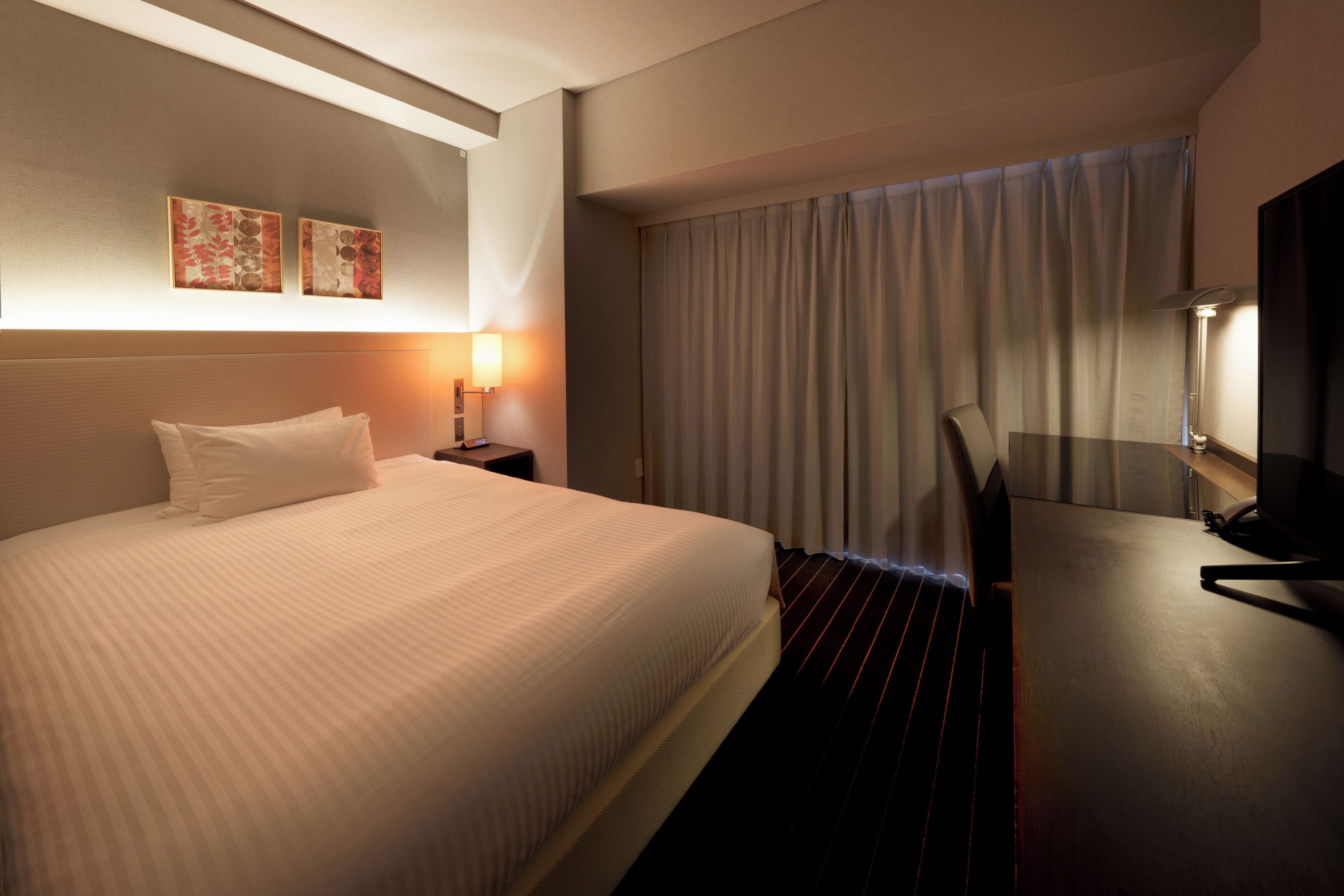 A hotel room with a large bed, a desk, a lamp, and two wall-mounted artworks.