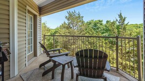 A covered balcony with two Adirondack chairs and a table, overlooking a forested area.