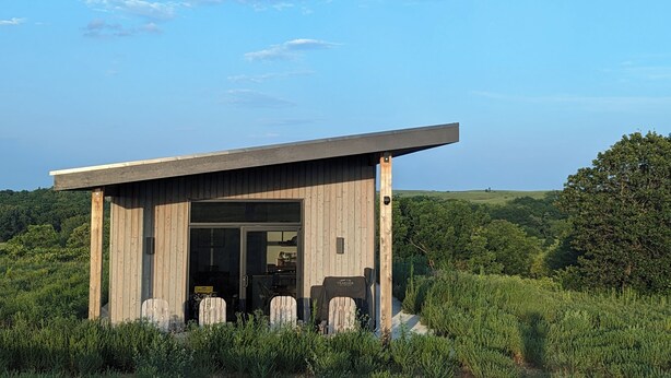 Eine Holzhütte mit einer großen Glasdoor, umgeben von Grün und blauem Himmel.