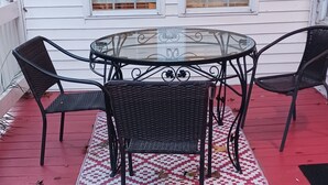 Une table à plateau en verre, entourée de quatre chaises en osier, est placée sur une terrasse rouge, elle-même bordée d’un tapis à motifs.