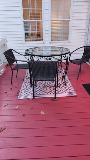 A glass-top table with four wicker chairs on a red deck, surrounded by a patterned rug.