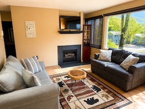Ein Wohnzimmer mit Kamin, zwei Sofas, einem Couchtisch und einem großen Fenster mit Blick auf Bäume.