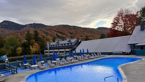 Piscina con sillas de descanso y sombrillas, rodeada por un edificio con montañas al fondo.