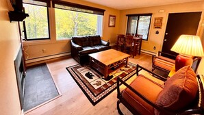 Una sala de estar con sofá, mesa de centro y sillones, una chimenea y ventanas amplias con vistas a árboles.