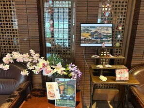 Una habitación japonesa tradicional con suelo de tatami, una mesa baja con un jarrón de flores, un televisor mostrando un paisaje y un cartel con texto en japonés.