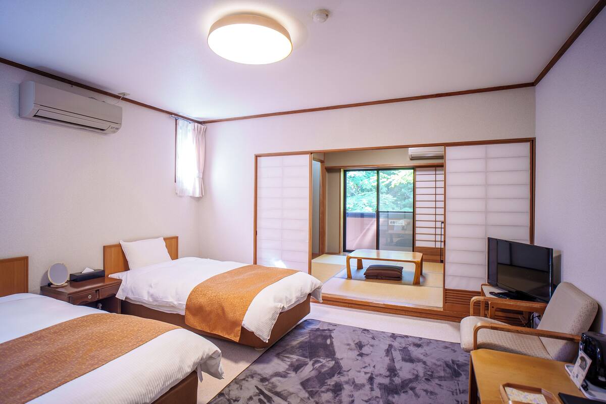 Habitación de hotel con dos camas, televisión, zona de estar y puerta corrediza que da a un balcón.