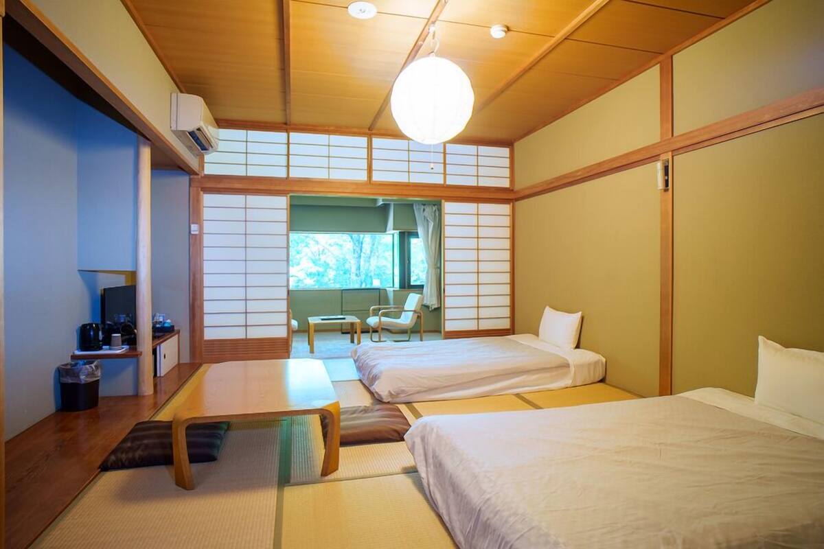 Una habitación tradicional japonesa con suelo de tatami, dos camas, una mesita y vistas a zonas verdes.