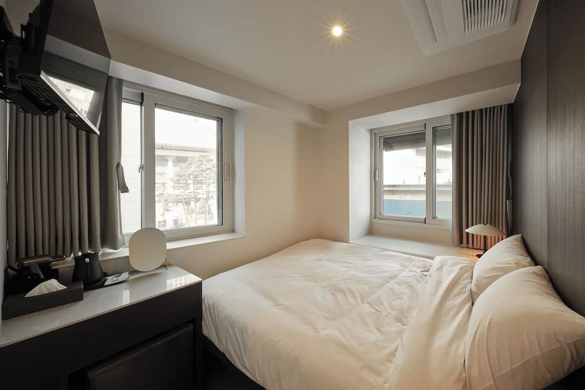 Une chambre d’hôtel avec un lit, un bureau avec une lampe, une fenêtre avec des rideaux et une vue sur l’extérieur.