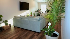 A living room with a grey sofa, wooden flooring, a flat-screen TV mounted on the wall, and several potted plants. - Masters Week Rental • 11 Miles to Augusta National • Masters Week Ready (Augusta)