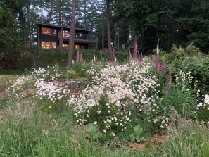 Ein Haus, umgeben von einem Garten mit weißen und rosafarbenen Blumen, hohen Bäumen und einer Steinmauer.
