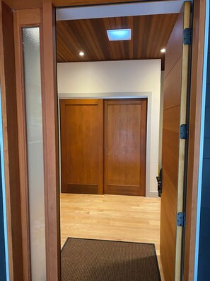 Eine Holztür mit einer strukturierten Fußmatte am Eingang, eine Decke mit eingelassener Beleuchtung und ein Blick in einen Raum mit Holzboden.