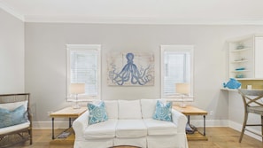 Une pièce avec un canapé blanc, deux tables d’appoint avec des lampes, une œuvre d’art représentant une pieuvre accrochée au mur, et une chaise en bois.