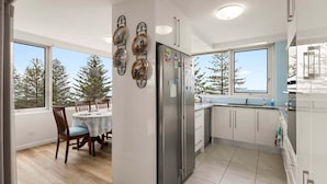 A modern kitchen with a dining area, large windows, and a view of trees. - Coastal Comfort with Ocean Views | Oceania 19 (Gold Coast)