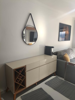 Quarto moderno com design minimalista, apresentando um sofá, uma mesa de centro, um aparador com espaço para vinhos, um espelho e uma moldura com uma imagem na parede.