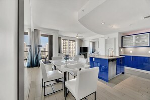 Une cuisine moderne avec des meubles bleus, un coin repas avec des chaises blanches et une grande fenêtre avec des rideaux.