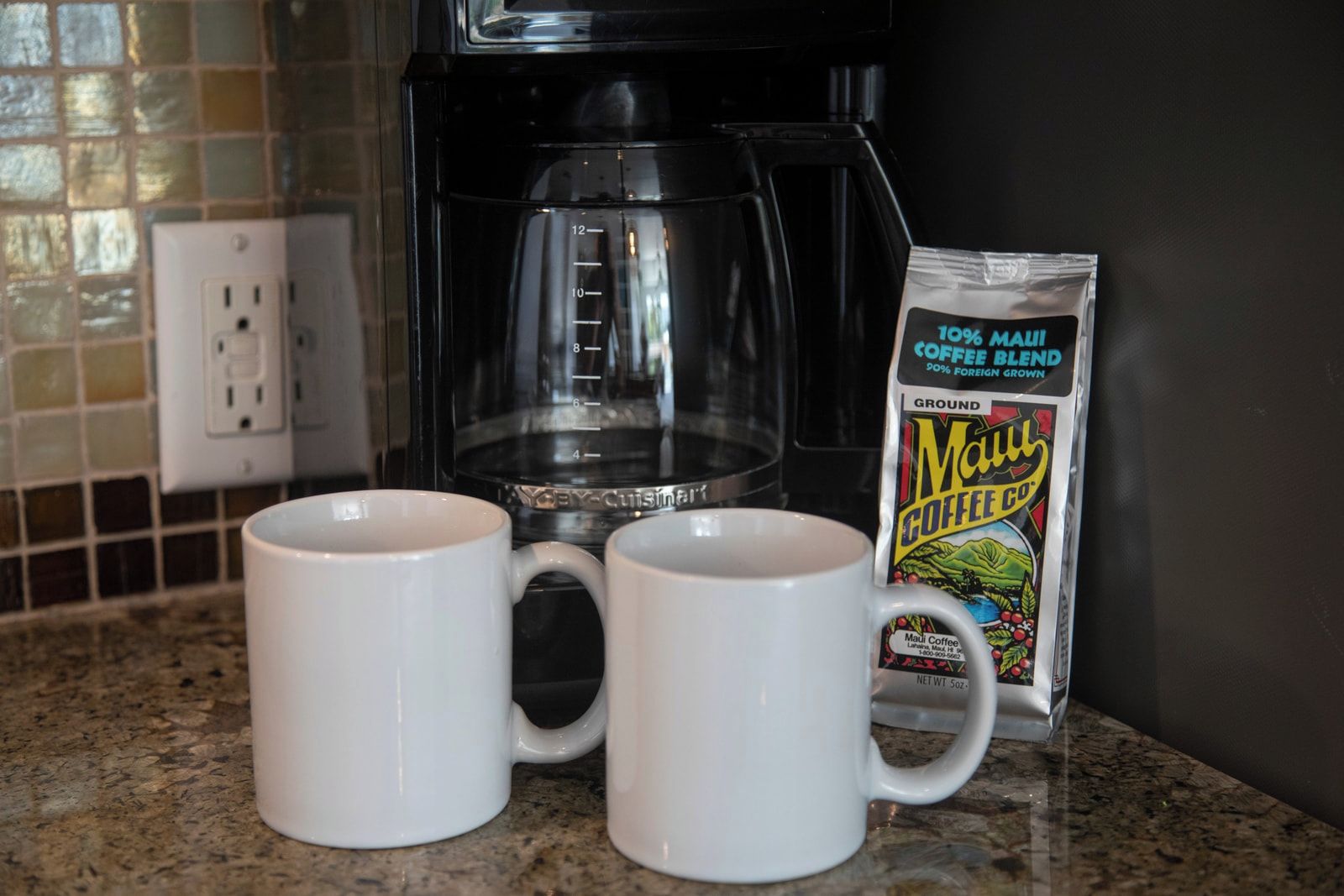 Dos tazas blancas sobre una encimera de cocina, una cafetera y un paquete de Café Maui.