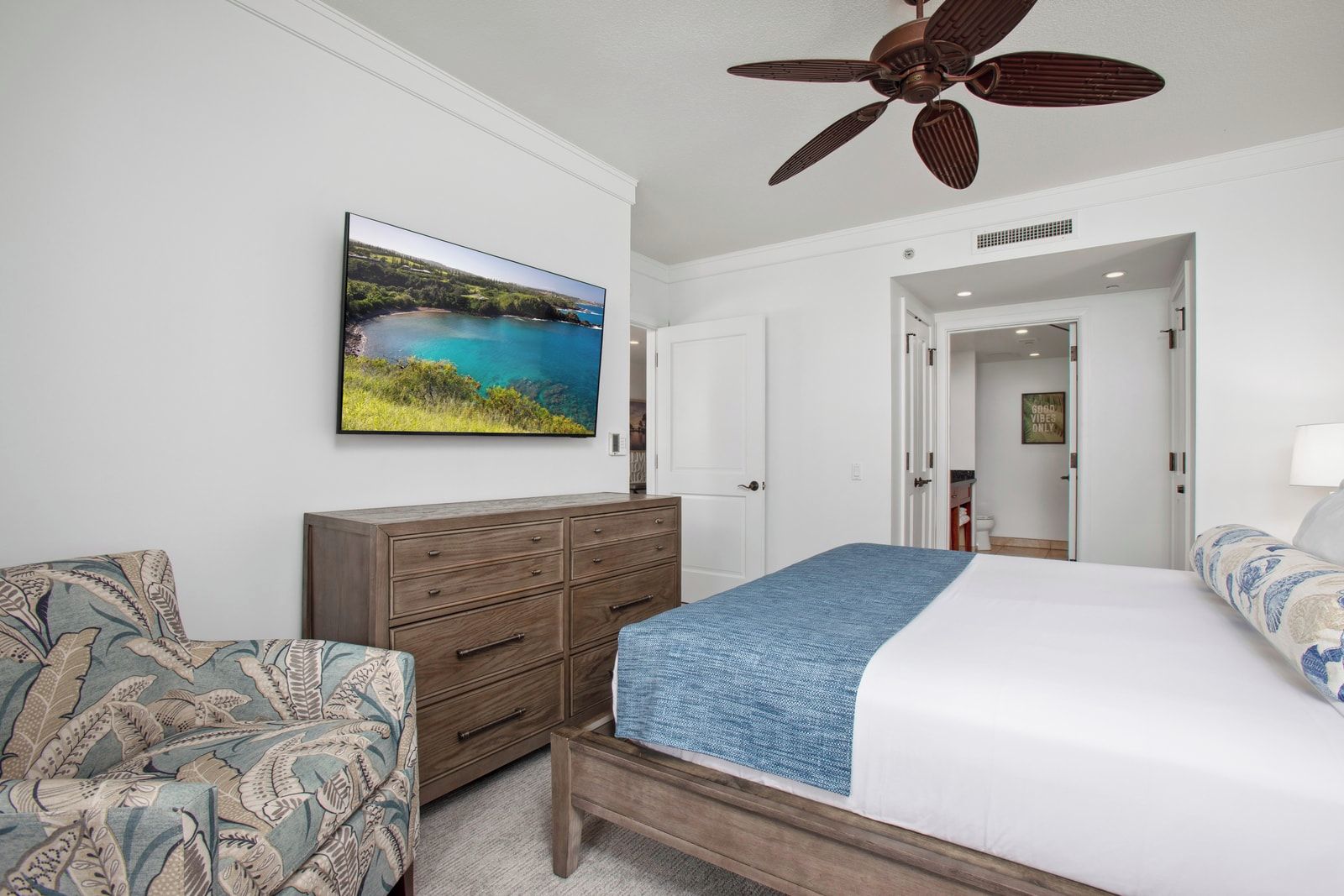 Une chambre à coucher comprenant un lit, une commode, une chaise, un ventilateur de plafond et un téléviseur diffusant un paysage pittoresque.