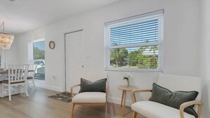 Una habitación con una mesa de comedor, dos sillas, un sofá, una mesita auxiliar con un florero y una ventana con persianas.