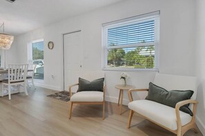 Ein Zimmer mit Esstisch, zwei Stühlen, einer Couch, einem kleinen Beistelltisch mit einer Vase und einem Fenster mit Jalousien.