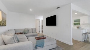 Una sala de estar moderna con un sofá, un televisor de pantalla plana montado en la pared y una cocina al fondo.