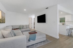 Ein modernes Wohnzimmer mit einer Couch, einem am Wand montierten Flachbildfernseher und einer Küchenzeile im Hintergrund.