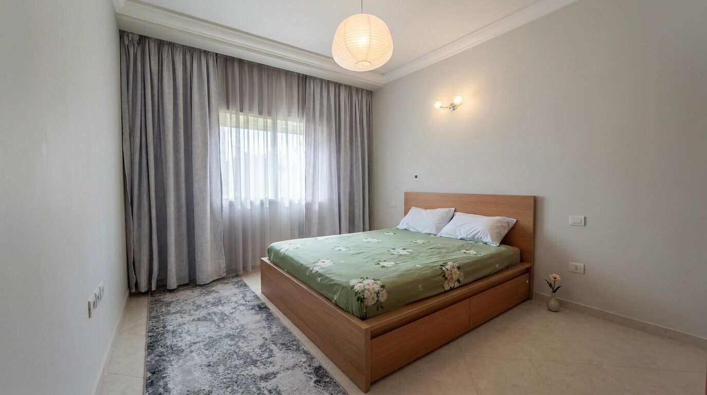 Un dormitorio con cama de madera, un ventanal con cortinas, una lámpara de pared y un jarrón pequeño con flores.
