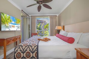 A hotel room with a bed, a TV, a ceiling fan, a window with curtains, and a view of palm trees and buildings. - 2 Honua Kai Villas | Ocean View | Sleeps 10 | Free Car w/ 6+ Nights | ML-2831 (Lahaina)
