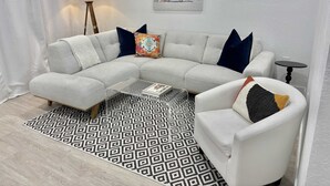 En moderne stue med en hjørnesofa, en hvid lænestol, et sofabord, et farverigt tæppe, en lampe og et indrammet billede på væggen.