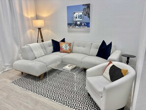 Ein modernes Wohnzimmer mit einer Eckcouch, einem weißen Sessel, einem Couchtisch, einem Teppich mit Muster und einer Lampe. An der Wand hängt ein gerahmtes Bild.
