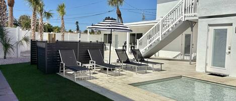 Un área de piscina moderna con sillas de descanso, una zona de estar techada y una escalera que conduce a un nivel superior.