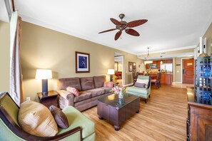 Ein Wohnzimmer mit einer Couch, Sesseln, einem Couchtisch, einem Deckenventilator und einer Küche im Hintergrund.