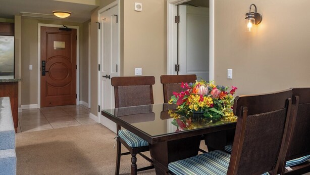 Un comedor con una mesa de vidrio, sillas acolchadas, un centro de mesa floral y una lámpara de pared.