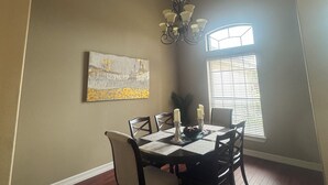 Een eetruimte met een tafel gedekt voor vier personen, een kroonluchter, een raam met jaloezieën en een schilderij aan de muur.