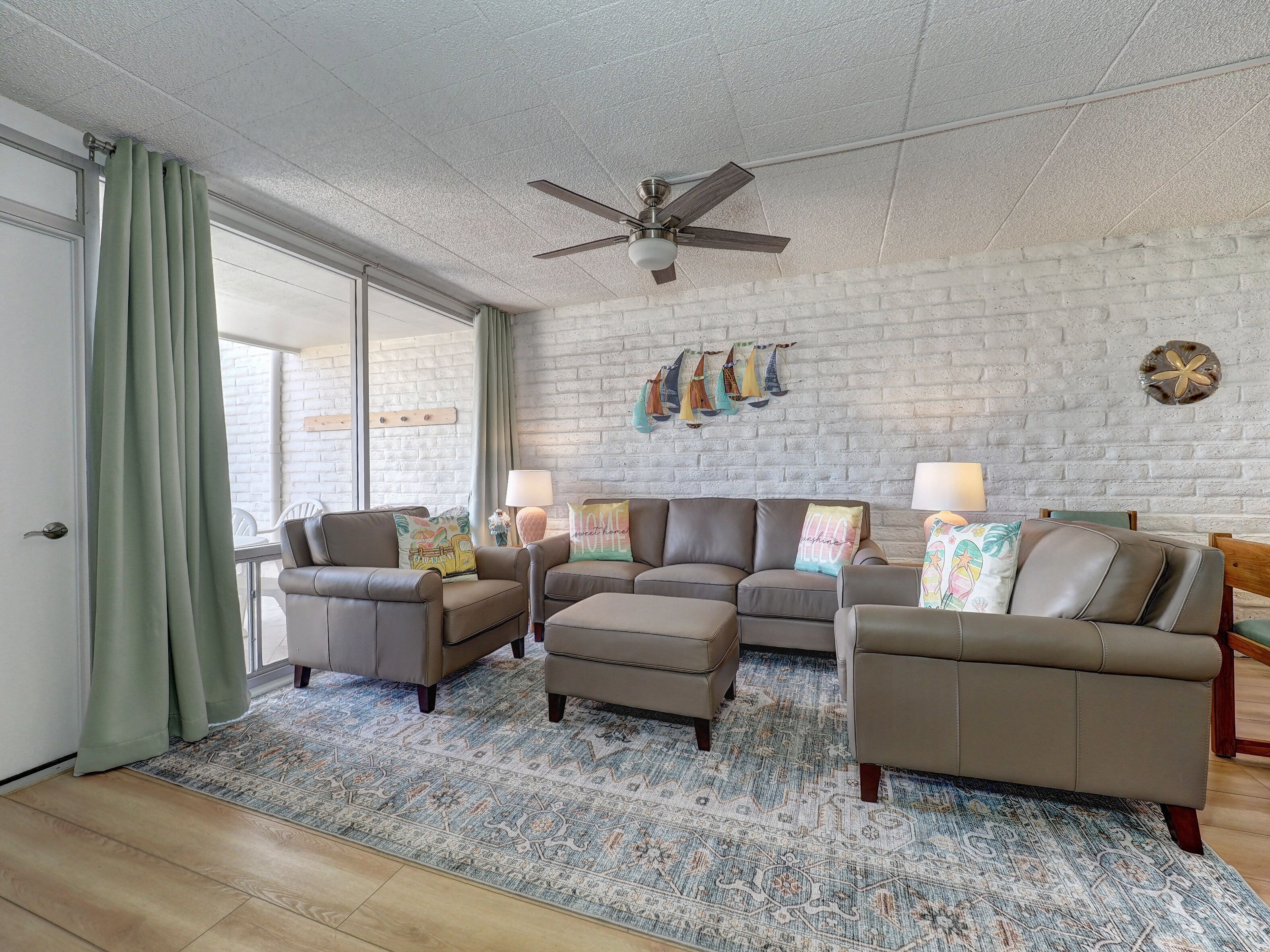 A living room with a sectional sofa, two armchairs, a ceiling fan, a patterned rug, and a brick wall with decorative pillows.