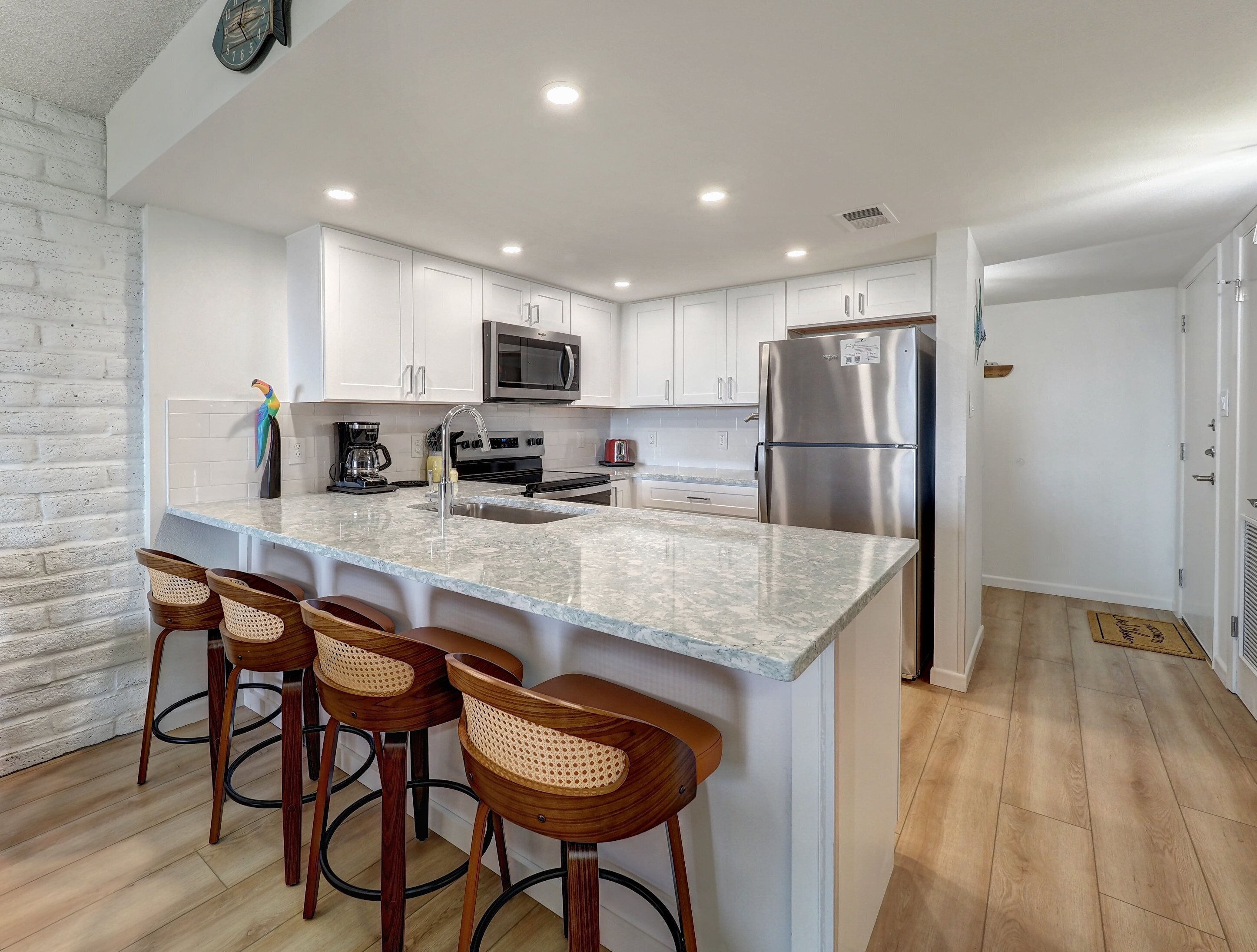 A modern kitchen with white cabinets, a stainless steel refrigerator, a microwave, and a coffee maker. There are three wooden bar stools with woven seats.