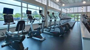 Ett gym med konditionsmaskiner, stora fönster och utsikt över parkerade bilar utanför.