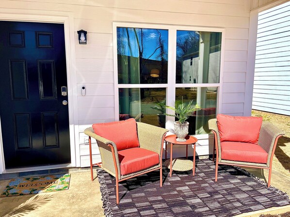 A front door with a welcome mat, two wicker chairs with red cushions, a small table with a vase, and a large window. - Sparkle City Vibes await for you to relax and enjoy! (Spartanburg)